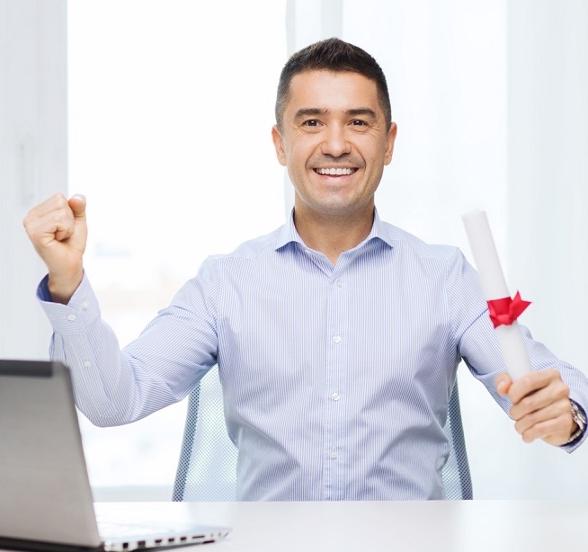 Homme assis à un bureau, tenant un diplôme roulé noué d’un ruban rouge tout en levant le poing. Un ordinateur portable est posé devant lui.