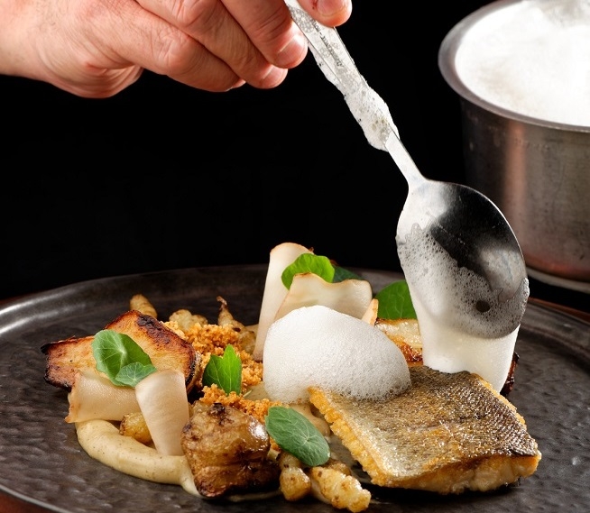 Gros plan sur une assiette de gastronomie contemporaine. Une main tient une cuillère au-dessus d'une préparation composée de poisson, mousses, légumes et éléments décoratifs.
