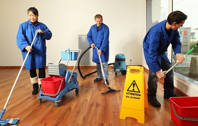 Image montrant trois personnes en blouse de travail bleue réalisant des opérations de nettoyage dans une pièce. L'une utilise un balai plat, une autre un aspirateur et la troisième nettoie une vitre. Au premier plan, un panneau jaune de signalisation indique « Attention sol glissant ».