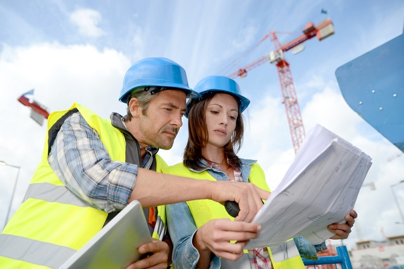 Un homme et une femme portant des casques de chantier bleus et des gilets de sécurité jaunes consultent des plans sur un chantier de construction. Une grue est visible à l'arrière-plan.