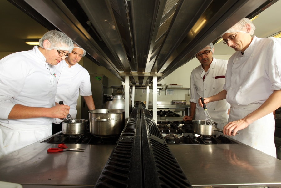 Quatre hommes en tenue de cuisine blanche et charlotte travaillent autour de plaques de cuisson dans une cuisine professionnelle.