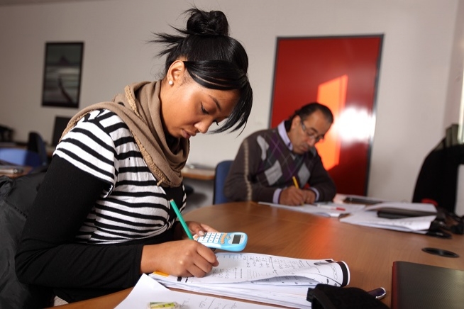 Femme écrivant sur une feuille à l'aide d'un crayon tout en utilisant une calculatrice. Une autre personne travaille sur des documents en arrière-plan.