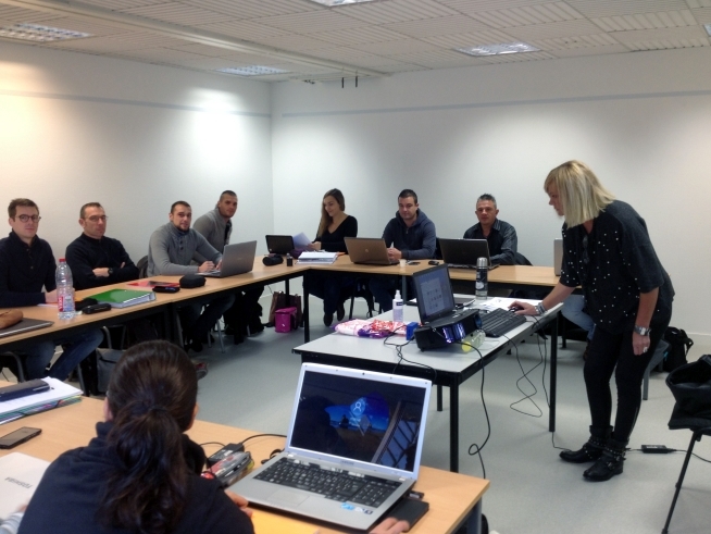 Personnes assises autour de tables disposées en U dans une salle de formation équipée d’ordinateurs portables.