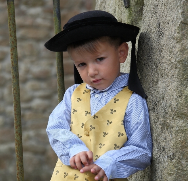 Petit garçon en costume traditionnel breton appuyé contre un mur en pierre, portant un chapeau noir, une chemise rayée et un gilet jaune à motifs.