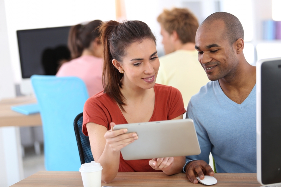 Une femme et un homme assis devant un ordinateur consultent une tablette numérique dans une salle informatique.