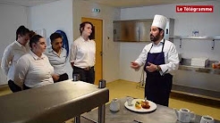 Homme en tenue de cuisine blanche et toque de chef présentant une assiette à un groupe de stagiaires dans une cuisine pédagogique.