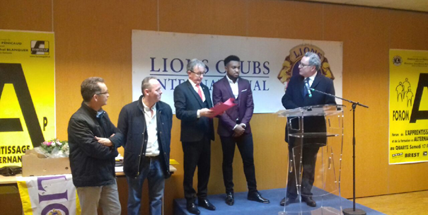 Plusieurs personnes se tiennent sur une scène lors d'une cérémonie de remise de prix. Une personne reçoit un document tandis qu'une autre s'exprime derrière un pupitre. Des affiches « Lions Clubs International » et « Foromap » sont visibles en arrière-plan.