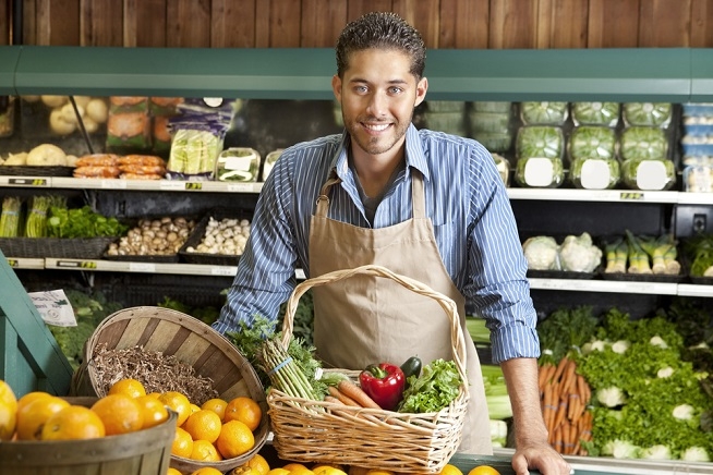 Homme portant un tablier derrière un étal de fruits et légumes frais dans un magasin. Un panier rempli de légumes est posé devant lui.