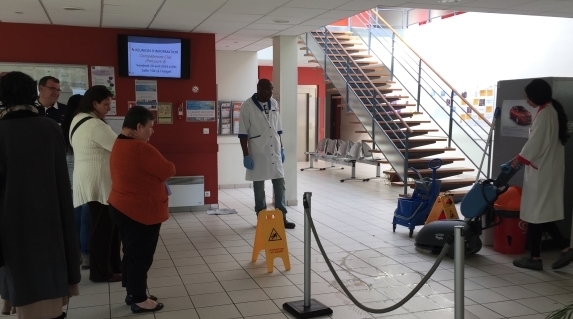 Plusieurs personnes observent une démonstration de matériel de nettoyage dans un hall. Une autolaveuse et différents équipements d'entretien sont utilisés dans un espace balisé.