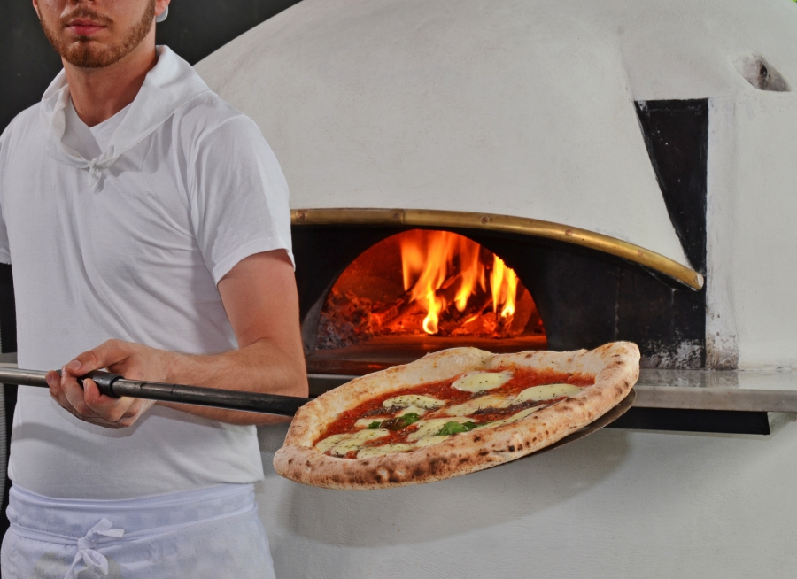 Homme en tenue de cuisine blanche tenant une pizza sur une pelle à pizza devant un four traditionnel allumé.