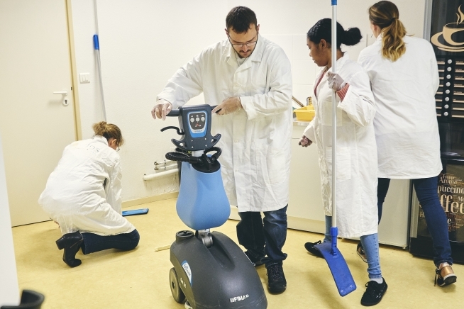 Personnes en blouse blanche suivant une démonstration autour d'une autolaveuse dans une salle de formation. Du matériel de nettoyage est visible au premier plan.