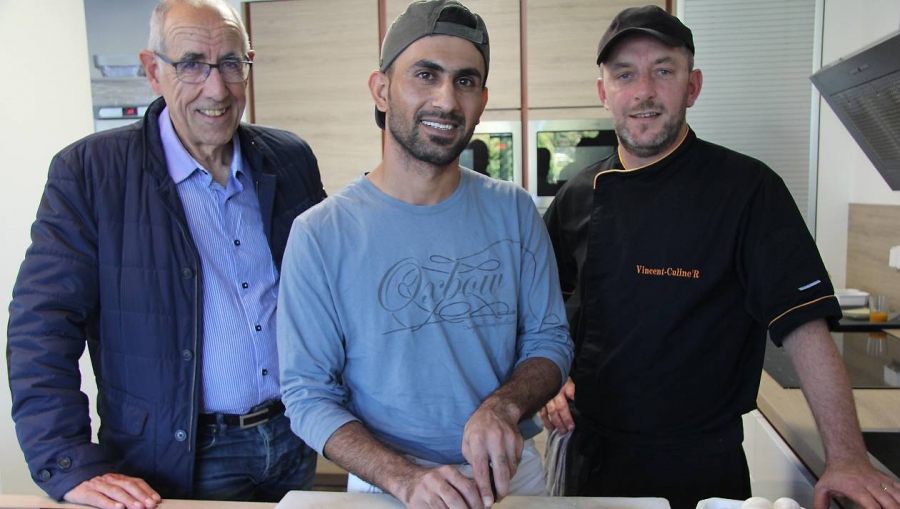 Trois hommes se tiennent côte à côte dans une cuisine professionnelle. Au centre, l'un est en tenue de travail et prépare des aliments sur un plan de travail tandis qu'un autre porte une veste de cuisinier noire.