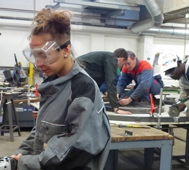 Personne portant des lunettes de protection et une tenue de travail dans un atelier technique, utilisant un outil sur un établi. Plusieurs autres personnes travaillent en arrière-plan autour de postes équipés de matériel de métallurgie et de fabrication.