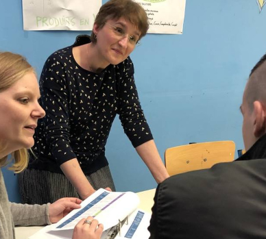 Trois personnes participent à un échange autour d'une table dans une salle de formation. Une femme est debout tandis que deux autres personnes sont assises avec des documents devant elles.