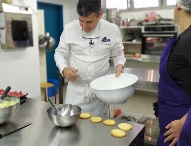 Homme en tenue de cuisine blanche tenant un grand saladier dans une cuisine professionnelle. Plusieurs fonds de tartelettes sont alignés sur un plan de travail en inox.