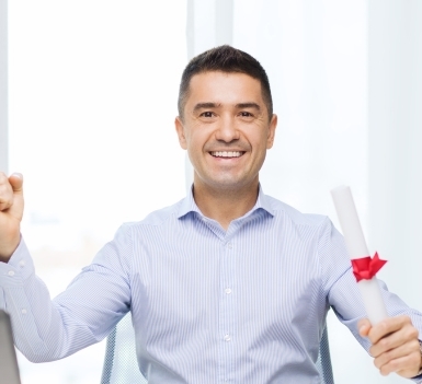 Homme assis dans un bureau tenant un diplôme roulé et attaché par un ruban rouge. Un ordinateur portable est partiellement visible sur la gauche.