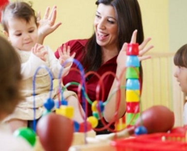 Femme assise à une table de jeu avec plusieurs jeunes enfants. Des jouets colorés d'éveil sont disposés au premier plan.