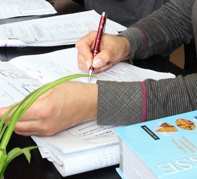 Main tenant un stylo rouge et écrivant sur des documents posés sur une table. Des cahiers, des feuilles de travail et des supports pédagogiques sont visibles.