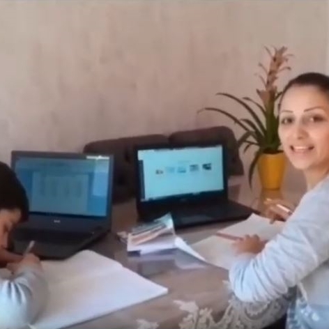 Femme assise à une table de travail à domicile avec un enfant. Deux ordinateurs portables ouverts, des cahiers et des supports de cours sont visibles sur la table.