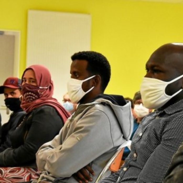 Photographie prise dans une salle aux murs jaune vif montrant plusieurs personnes assises côte à côte sur des chaises. Au premier plan, trois personnes portent des masques de protection blancs ou à motifs. Une personne porte une veste à capuche grise, une autre une chemise sombre à petits motifs et une troisième un foulard rose couvrant les cheveux. D'autres participants masqués sont visibles à l'arrière-plan.