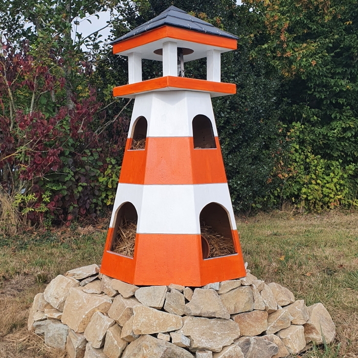 Photographie en extérieur d'une structure en bois en forme de phare installée sur un socle circulaire composé de pierres claires. Le phare présente des bandes horizontales orange vif et blanches, quatre ouvertures en arc de cercle dans sa partie inférieure et un lanternon carré surmonté d'un toit gris foncé. L'ensemble est placé sur une pelouse devant une haie dense composée d'arbustes verts, rouges et bruns.