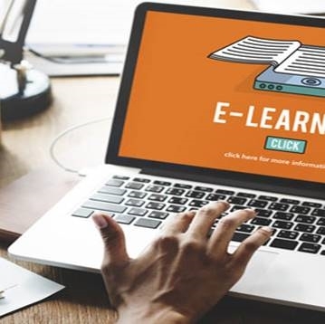 Photographie en gros plan d’un ordinateur portable ouvert posé sur un bureau en bois. Une main est placée sur le clavier et le pavé tactile de l’ordinateur. L’écran affiche une interface orange avec l’inscription « E‑LEARNING », un bouton « CLICK » et une illustration représentant des documents numériques. Une lampe de bureau et d’autres éléments de travail sont partiellement visibles en arrière-plan.
