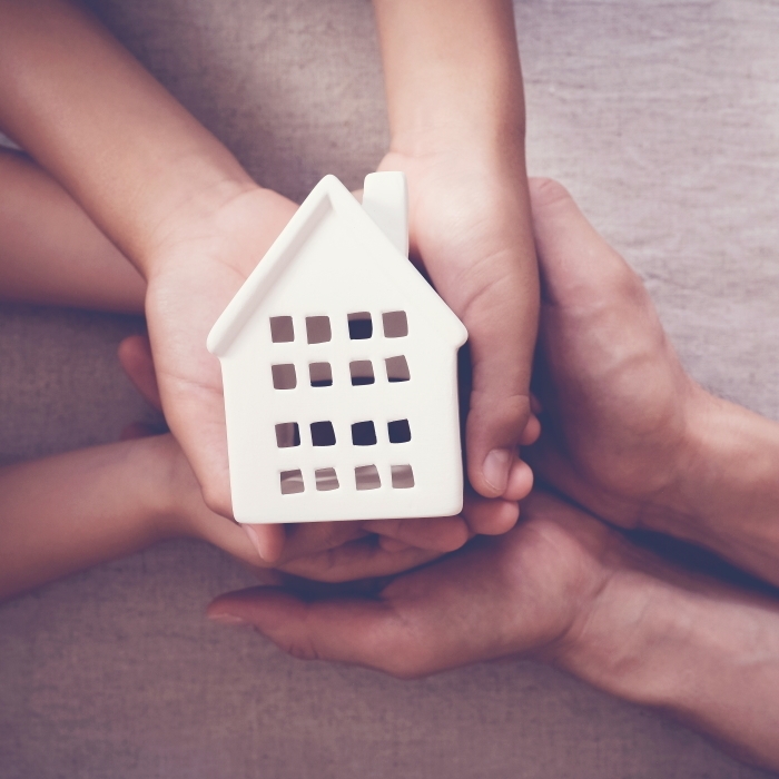 Plusieurs mains entourent et soutiennent une petite maison blanche miniature posée au centre de l'image. Les mains sont disposées en cercle sur un fond beige et mettent en valeur la maquette représentant une habitation.