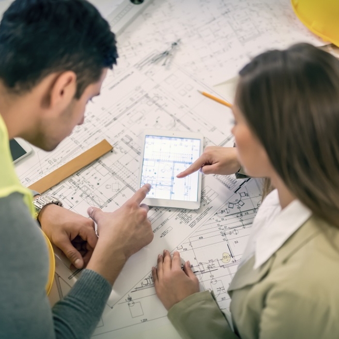 Un homme à gauche et une femme à droite consultent une tablette numérique affichant un plan technique, au-dessus de plans d’architecture étalés sur une table. Une règle, un crayon et un casque de chantier sont visibles autour des documents.