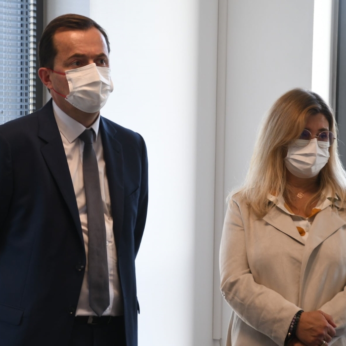 Deux personnes portant des masques de protection se tiennent debout dans une pièce lumineuse aux murs blancs. Une personne est vêtue d'un costume sombre avec une cravate et l'autre porte une veste claire et des lunettes.