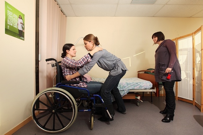 Femme assise dans un fauteuil roulant tandis qu'une autre femme l'aide à se relever ou à effectuer un transfert. Une troisième femme observe la scène dans une chambre ou une salle pédagogique.