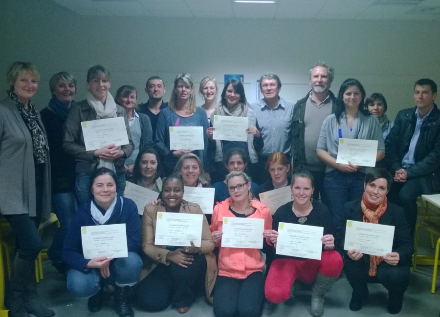 Groupe d'une vingtaine de personnes posant dans une salle de formation. Plusieurs participants tiennent un diplôme ou un certificat devant eux.
